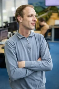 Justdata ICT, Een man met een lichtblauwe trui met kwart rits staat met zijn armen over elkaar, glimlacht en kijkt opzij in een modern kantoor met computers en een andere persoon aan het werk op de achtergrond.