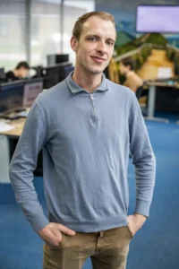 Justdata ICT, Een man in een lichtblauwe trui en beige broek staat in een kantoor met zijn handen in zijn zakken en lacht naar de camera. Op de achtergrond werken verschillende mensen aan computertafels.