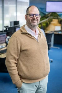 Justdata ICT, Een man met een bril en een baard, in een bruine trui en lichte broek, staat glimlachend met zijn hand in zijn zak in een kantoor met computertafels en monitoren op de achtergrond.