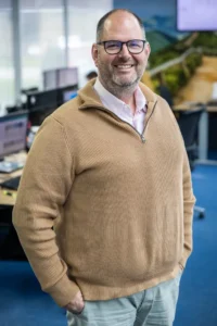 Justdata ICT, Een lachende man met bril en baard, die een bruine trui draagt over een overhemd met kraag, staat met zijn handen in zijn zakken in een modern kantoor met bureaus en computerschermen op de achtergrond.