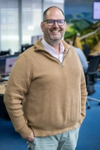 Justdata ICT, Een lachende man met bril en baard draagt een bruine trui en staat in een kantoor met computers, bureaus en blauwe vloerbedekking.