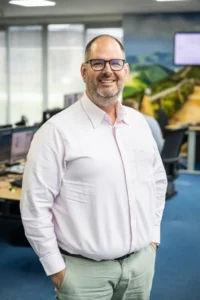 Justdata ICT, Een man met een bril, een korte baard en kort haar staat glimlachend in een kantoor met bureaus, computers en een muurschildering van een landweggetje op de achtergrond. Hij draagt een lichtroze overhemd en een grijze broek.