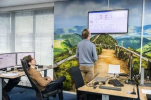 Justdata ICT, Twee mensen in een kantoor kijken naar een groot scherm met datadashboards. Op de bureaus staan meerdere monitoren en computers en op de muur achter hen is een schilderachtig bergpad afgebeeld.