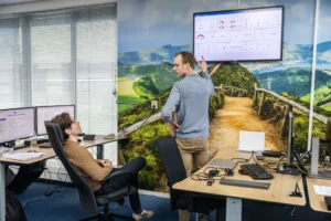 Justdata ICT, Twee mannen in een kantoor bespreken gegevens die worden weergegeven op een groot scherm aan de muur; de een zit aan een bureau met meerdere monitoren, terwijl de ander staat en naar het scherm gebaart. Een muurschildering in de natuur bedekt de achterwand.