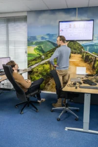 Justdata ICT, Twee mensen in een kantoor bespreken gegevens die op een grote monitor aan de muur worden getoond. Eén persoon staat en wijst naar het scherm, terwijl de ander in een stoel zit. Op de muur van het kantoor is een schilderachtige muurschildering te zien van een groen pad door heuvels.