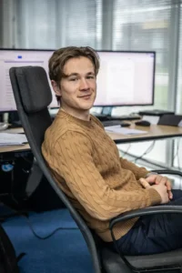 Justdata ICT, Een jonge man met kort bruin haar en een bruine kabeltrui zit in een bureaustoel en lacht naar de camera. Achter hem staan grote computerschermen en een bureau met papieren.