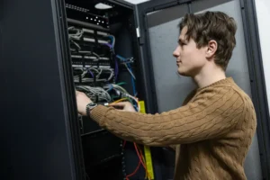 Justdata ICT, Een persoon met een bruine trui werkt met netwerkkabels in een serverrack, waarschijnlijk voor onderhoud of probleemoplossing in een serverruimte.