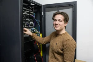 Justdata ICT, Een persoon in een bruine trui staat naast een open serverrack en lacht terwijl hij werkt met netwerkkabels en apparatuur in de serverkast.