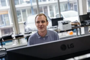 Justdata ICT, Een man in een lichtgrijs zip-up shirt zit en glimlacht voor een computermonitor in een helder kantoor met grote ramen en verschillende kantoorapparatuur op de achtergrond.