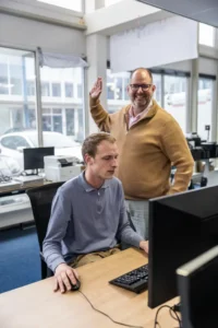 Justdata ICT, Een man zit aan een bureau en gebruikt een computer terwijl een andere man naast hem zijn hand opsteekt en lacht in een kantoor met grote ramen en meerdere beeldschermen.
