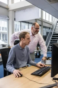 Justdata ICT, Twee mannen in een kantoor, de een zit en gebruikt een computer terwijl de ander achteraan staat, glimlacht en naar het beeldscherm kijkt. Ze lijken samen te werken aan een taak in een moderne werkruimte.