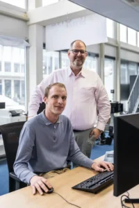 Justdata ICT, Twee mannen in een kantooromgeving: de een zit achter een bureau en gebruikt een computer, de ander staat naast hem en glimlacht naar de camera. Op de achtergrond zijn grote ramen en kantoorapparatuur te zien.