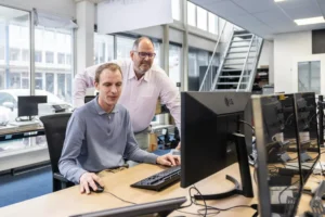 Justdata ICT, Twee mannen in een kantoor, de een zittend en met een computer, de ander staand naast hem en glimlachend. Ze worden omringd door bureaus, beeldschermen en grote ramen met daglicht.