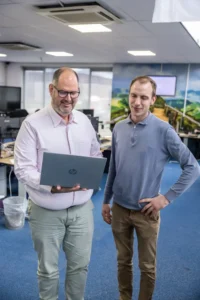 Justdata ICT, Twee mannen staan in een kantoor en kijken naar een laptop die één van hen vasthoudt. Beiden glimlachen en op de achtergrond zijn bureaus met computers en kantoorapparatuur zichtbaar.