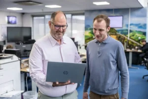 Justdata ICT, Twee mannen staan in een kantoor, kijken samen naar een laptopscherm en glimlachen. De achtergrond van het kantoor bestaat uit bureaus, computers en grote ramen met binnenvallend daglicht.