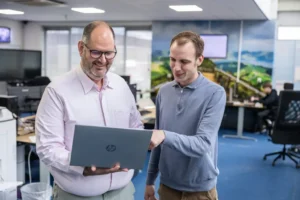 Justdata ICT, Twee mannen staan in een kantoor, glimlachen en kijken naar een laptop die één van hen vasthoudt. Eén man wijst naar het scherm van de laptop en op de achtergrond zijn bureaus met computers te zien.
