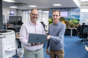Justdata ICT, Twee mannen staan in een modern kantoor, glimlachen en houden samen een open HP-laptop vast. Kantoorapparatuur en collega's zijn zichtbaar op de achtergrond.