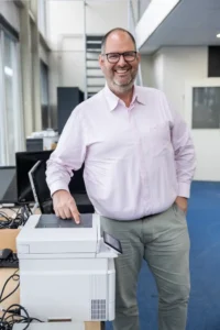 Justdata ICT, Een lachende man met een bril en een lichtroze overhemd staat naast een kantoorprinter en wijst ernaar met één hand terwijl de andere in zijn zak rust. Hij staat in een lichte, moderne kantooromgeving.