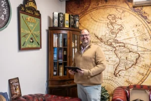 Justdata ICT, Een lachende man staat in een gezellige kamer met vintage inrichting en houdt een tablet vast. Achter hem hangen een grote antieke wereldkaart aan de muur, een houten kast met flessen en een retro-stijl snookerbord.