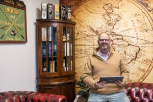 Justdata ICT, Een man met een bril en een bruine trui zit glimlachend op een roodleren bank en houdt een tablet vast. Achter hem hangt een muurschildering van een vintage landkaart, een houten kast met flessen en een bord met snooker op de muur.