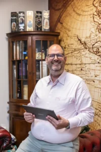 Justdata ICT, Een lachende man met bril en lichtroze overhemd houdt een tablet vast terwijl hij in een kantoor zit met een vintage wereldkaart aan de muur en een houten kast met boeken en flessen op de achtergrond.