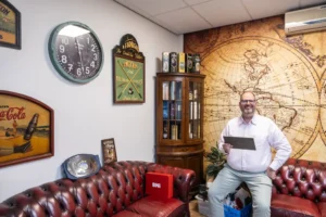 Justdata ICT, Een lachende man in een wit overhemd houdt een tablet vast terwijl hij op een roodleren bank zit in een kantoor dat is ingericht met vintage borden, klokken, een muurschildering van een landkaart en een glazen kast.
