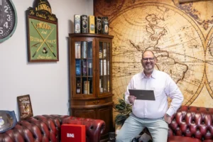 Justdata ICT, Een man met een wit overhemd en een bril zit op een roodleren bank, glimlacht en houdt een tablet vast. Achter hem staan vintage decorstukken, een houten kast met flessen en een grote muurschildering van een wereldkaart.