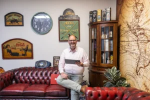 Justdata ICT, Een man met een roze overhemd en bril zit op een roodleren bank en houdt een tablet vast, omringd door vintage decor, waaronder wandklokken, uithangborden, een wereldbolkast en een grote kaart aan de muur.