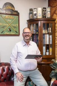 Justdata ICT, Een lachende man met bril en lichtroze overhemd zit op een roodleren stoel en houdt een tablet vast. Achter hem staan planken met flessen en verzamelobjecten, en aan de muur hangt een vintage bord met snooker en biljart.