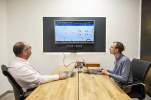 Justdata ICT, Twee mannen zitten aan een vergadertafel met laptops en kijken naar een groot scherm aan de muur met grafieken en datadashboards tijdens een vergadering in een modern kantoor.