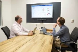 Justdata ICT, Twee mannen zitten aan een vergadertafel met laptops en kijken naar een groot scherm aan de muur waarop grafieken te zien zijn, wat doet denken aan een zakelijke bijeenkomst of een discussie over gegevensanalyse.