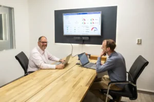 Justdata ICT, Twee mannen zitten tegenover elkaar aan een houten vergadertafel met laptops, glimlachen en praten. Een scherm aan de muur toont kleurrijke grafieken en datadashboards. De setting is een moderne vergaderruimte op kantoor.