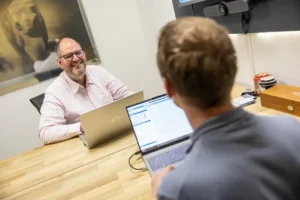 Justdata ICT, Twee mannen zitten tegenover elkaar aan een houten tafel met laptops en voeren een gesprek in een kantoor. Eén man glimlacht en op de achtergrond hangt een grote foto van een hond aan de muur.