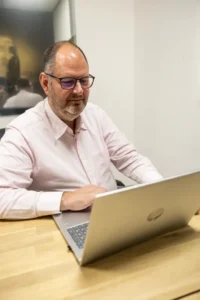 Justdata ICT, Een man met een bril en een baard, draagt een lichtroze overhemd en zit aan een houten bureau te werken op een HP laptop in een modern kantoor.