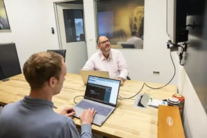 Justdata ICT, Twee mannen zitten aan een houten vergadertafel met laptops, de een kijkt naar een scherm aan de muur, de ander typt. Kantoorsetting, glazen kantoormuren en een afbeelding van een olifant zichtbaar op de achtergrond.