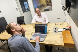 Justdata ICT, Twee mannen zitten aan een houten tafel in een kantoor, allebei met een laptop. De ene man kijkt peinzend op naar een scherm aan de muur, terwijl de andere glimlacht. Op de tafel liggen verschillende kantoorartikelen.