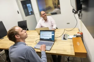 Justdata ICT, Twee mannen zitten aan een houten bureau in een kantoor en gebruiken allebei een laptop. De ene man kijkt naar een scherm aan de muur, terwijl de andere naar hem kijkt. Op de tafel staan ook bureaustoelen en een grote houten kist.