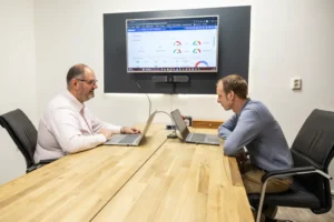Justdata ICT, Twee mannen zitten tegenover elkaar aan een houten vergadertafel en werken allebei op een laptop. Een scherm aan de muur achter hen toont grafieken en diagrammen met prestatiegegevens.
