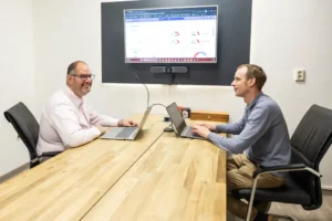 Justdata ICT, Twee mannen zitten aan een houten vergadertafel met laptops en hebben een vergadering. Een groot scherm achter hen toont grafieken en diagrammen. Beiden zijn bezig en glimlachen in een moderne kantooromgeving.