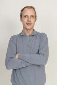 Justdata ICT, Een man met kort lichtbruin haar en een lichtgrijs shirt met lange mouwen staat tegen een effen lichte achtergrond met zijn armen over elkaar en een lichte glimlach op zijn gezicht.