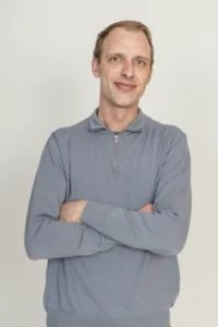 Justdata ICT, Een man met kort lichtbruin haar en een lichtgrijze trui met ritssluiting staat tegen een effen lichte achtergrond en glimlacht zachtjes met zijn armen over elkaar.