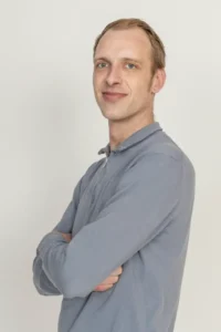 Justdata ICT, Een man met kort lichtbruin haar en blauwe ogen staat tegen een effen witte achtergrond, draagt een lichtgrijs shirt met lange mouwen, zijn armen gekruist en glimlacht lichtjes.