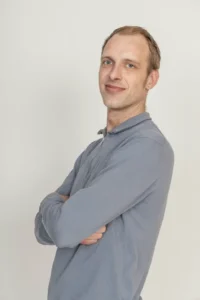 Justdata ICT, Een man met kort lichtbruin haar en blauwe ogen staat tegen een effen witte achtergrond, draagt een lichtblauw shirt met lange mouwen en glimlacht met zijn armen over elkaar.