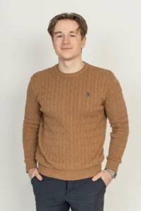 Justdata ICT, Een jonge man met kort lichtbruin haar staat te glimlachen tegen een effen achtergrond. Hij draagt een bruine, kabelgebreide trui en een donkere broek, met zijn handen in zijn zakken en een horloge om zijn linkerpols.