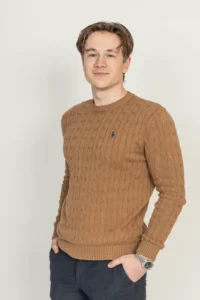 Justdata ICT, Een jonge man met kort bruin haar draagt een bruine kabeltrui en een donkere broek. Hij staat tegen een effen lichte achtergrond, glimlacht lichtjes, met zijn handen in zijn zakken.