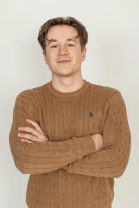 Justdata ICT, Een jonge man met kort bruin haar staat tegen een effen lichte achtergrond, draagt een bruine kabeltrui en glimlacht lichtjes met zijn armen over elkaar.