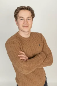 Justdata ICT, Een jonge man met een lichte huid en kort bruin haar staat glimlachend met zijn armen over elkaar en draagt een bruine kabeltrui tegen een effen lichte achtergrond.