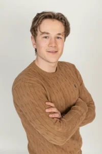 Justdata ICT, Een jonge man met lichtbruin haar die een bruine kabeltrui draagt, staat met zijn armen over elkaar en glimlacht lichtjes naar de camera tegen een effen lichte achtergrond.