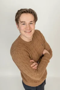 Justdata ICT, Een jonge man met lichtbruin haar die een bruine trui met structuur draagt, staat met zijn armen over elkaar en glimlacht naar de camera tegen een effen lichte achtergrond.