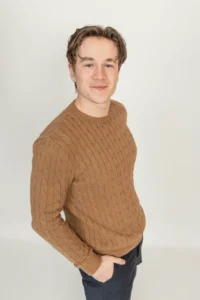 Justdata ICT, Een jonge man met een lichte huid en kort bruin haar staat zacht glimlachend tegen een effen lichte achtergrond. Hij draagt een bruine kabeltrui en een donkere broek, met één hand in zijn zak.